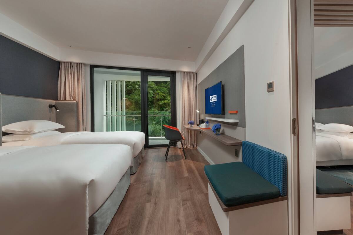 A modern hotel room with two beds, a desk, a TV, and a balcony with a view of greenery.