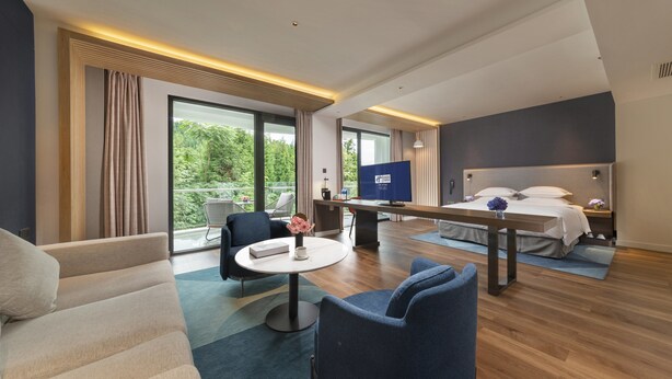 A hotel room with a bed, a desk, a chair, a TV, and a balcony with a view of greenery. - Holiday Inn Express E'Mei Qiliping by IHG (Meishan)
