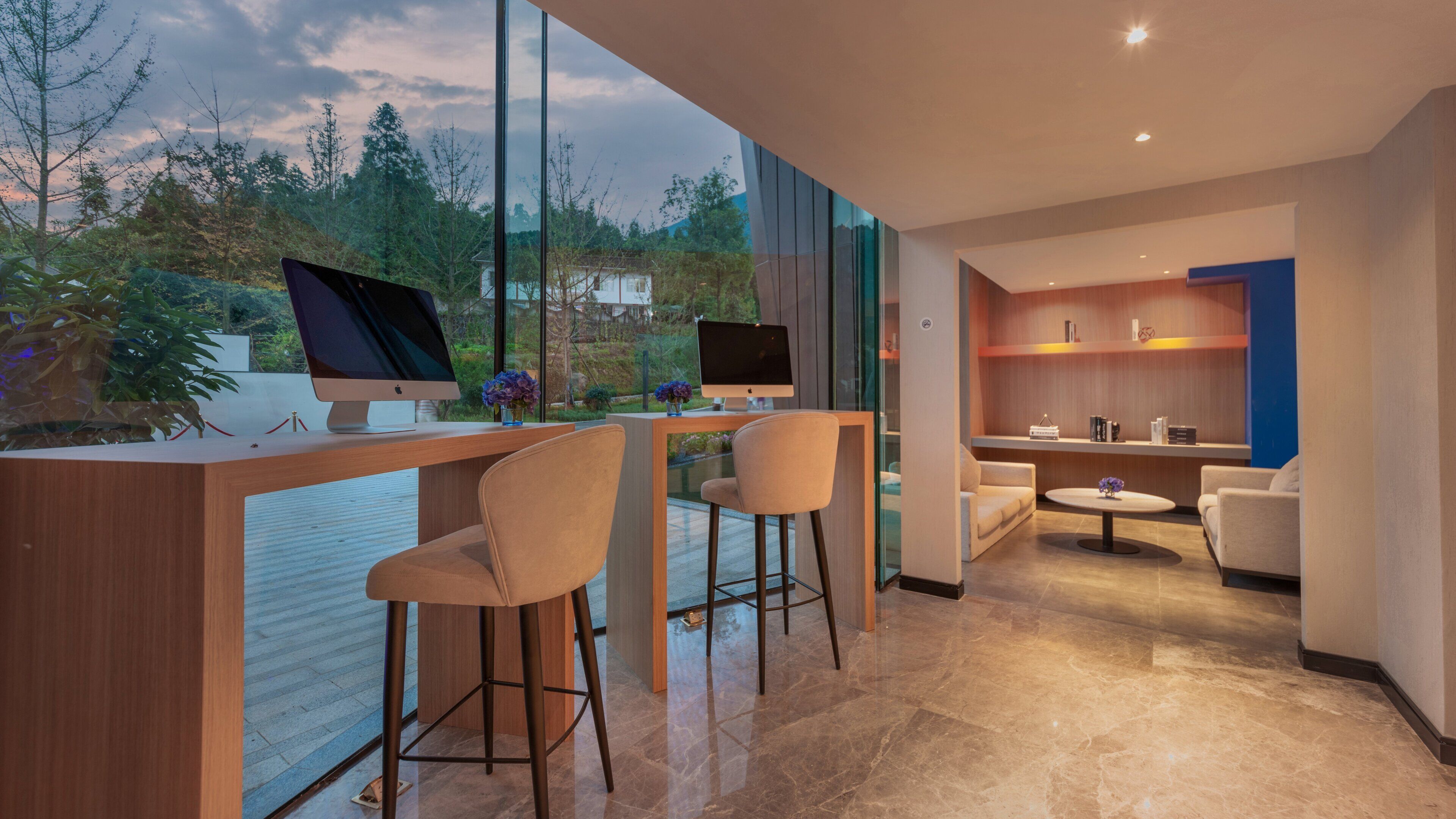 A modern interior with a desk, two chairs, a sofa, and a coffee table, with a view of trees and a building outside.