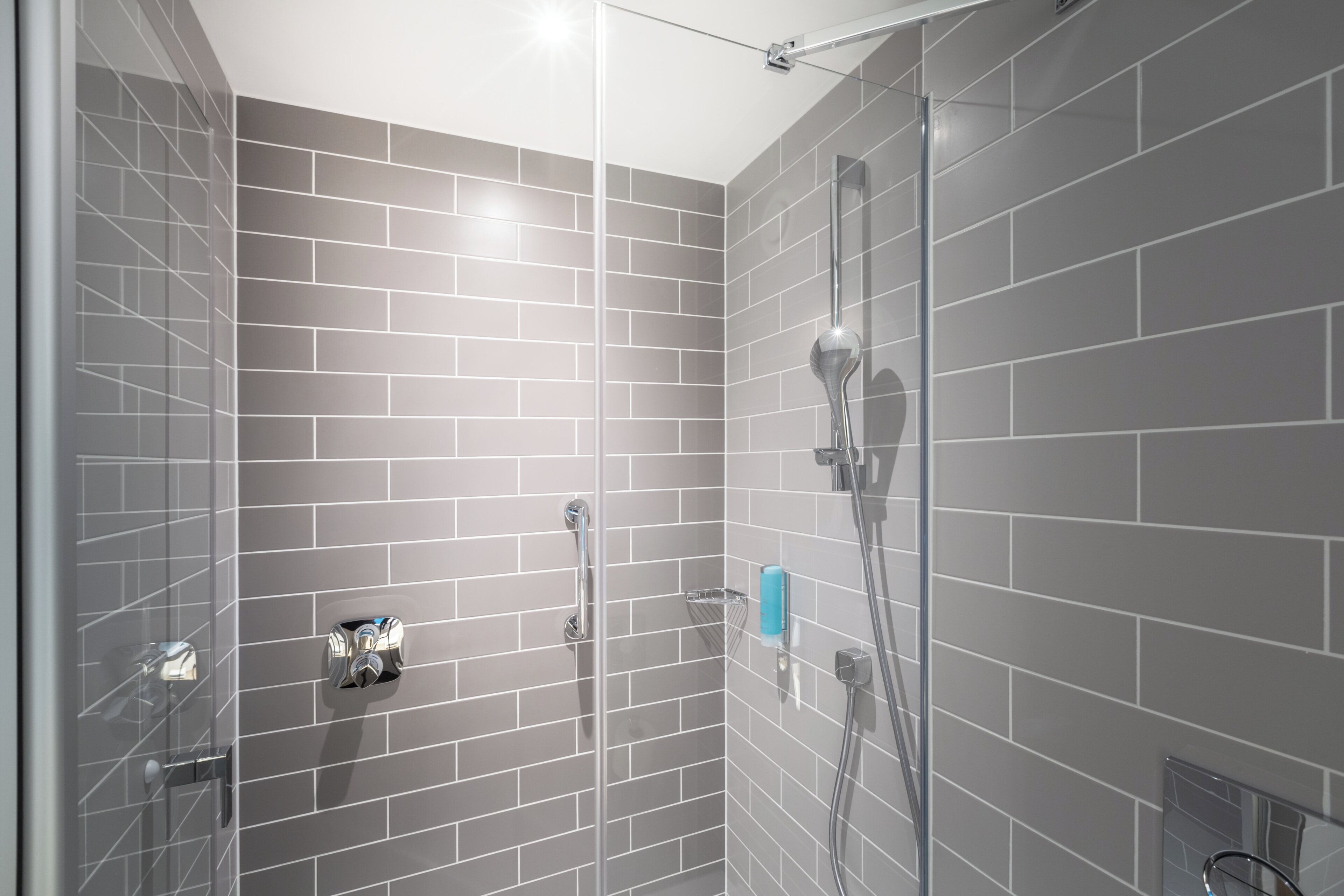 A shower with a glass door, grey tiled walls, and a handheld showerhead.