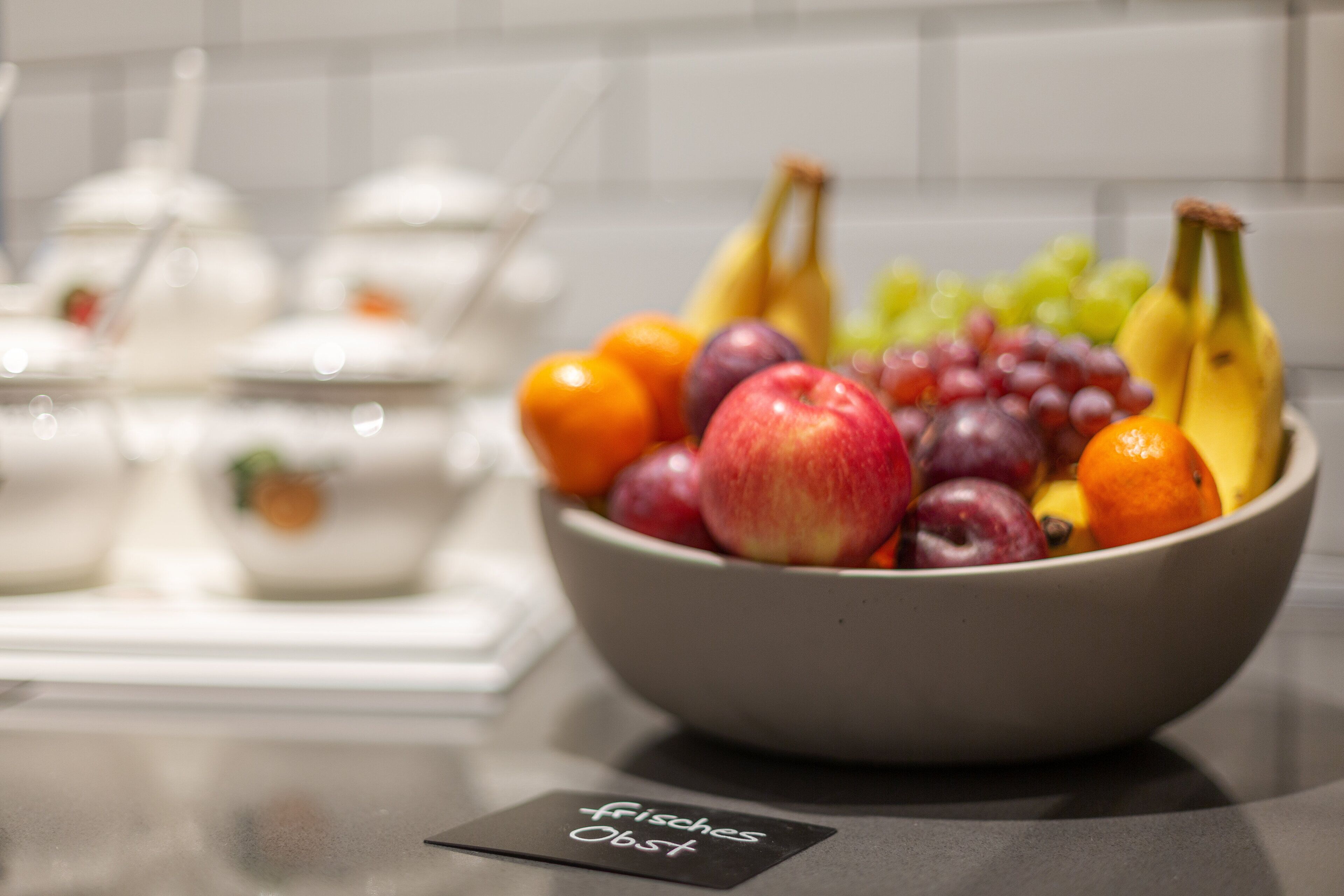 Un saladier rempli de fruits est posé sur une table, et une pancarte à côté indique : « Früchte von Fischers ».