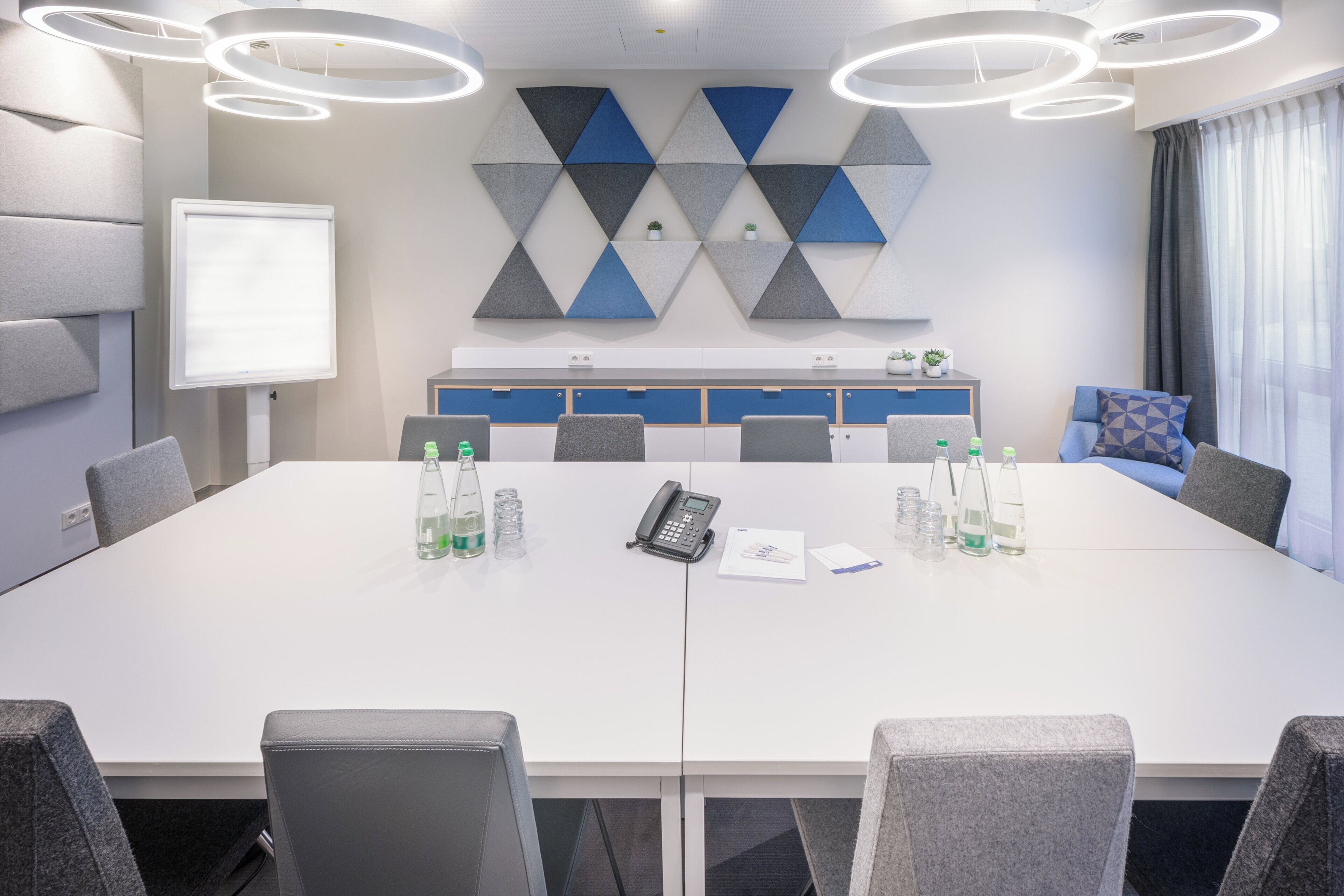 A conference room with a long table, chairs, a whiteboard, and a geometric wall decoration.