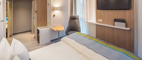 Una habitación de hotel con cama, televisor montado en la pared, escritorio y silla.