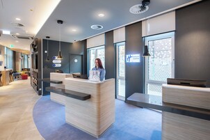 Área de recepção de hotel com design moderno, incluindo balcão de atendimento, espaço de estar e vista da cidade através de grandes janelas.