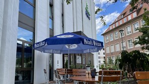 An outdoor seating area with tables and chairs, a blue umbrella with 'Maisel's Weisse' branding, and a building with large windows in the background.