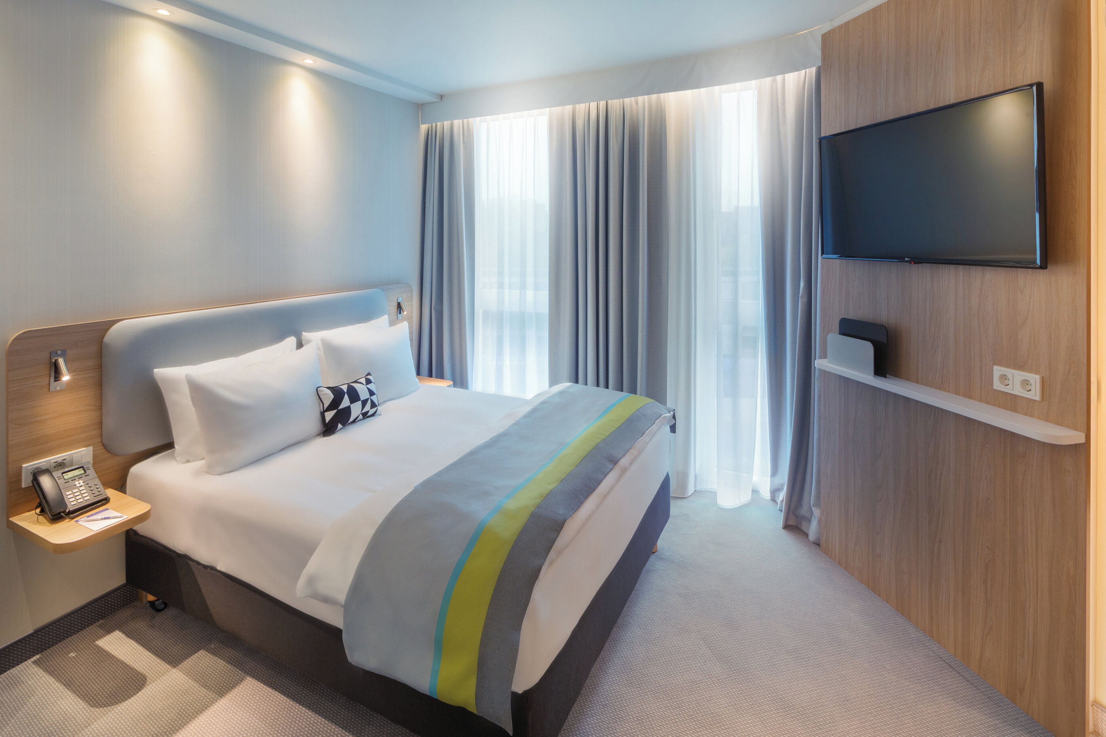 A hotel room with a bed, a TV mounted on the wall, a bedside table with a phone, and a window with curtains.