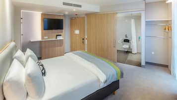A modern hotel room with a bed, a TV, a wardrobe, and a bathroom visible through an open door.