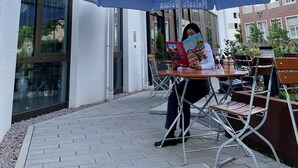 A person sitting at an outdoor table under a blue umbrella with the text "Maisel's Weisse" on it, in front of a building with large windows.
