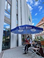 Uma pessoa sentada a uma mesa ao ar livre sob um guarda-sol azul com a inscrição "Maisel's Weisse", em frente a um prédio com grandes janelas.