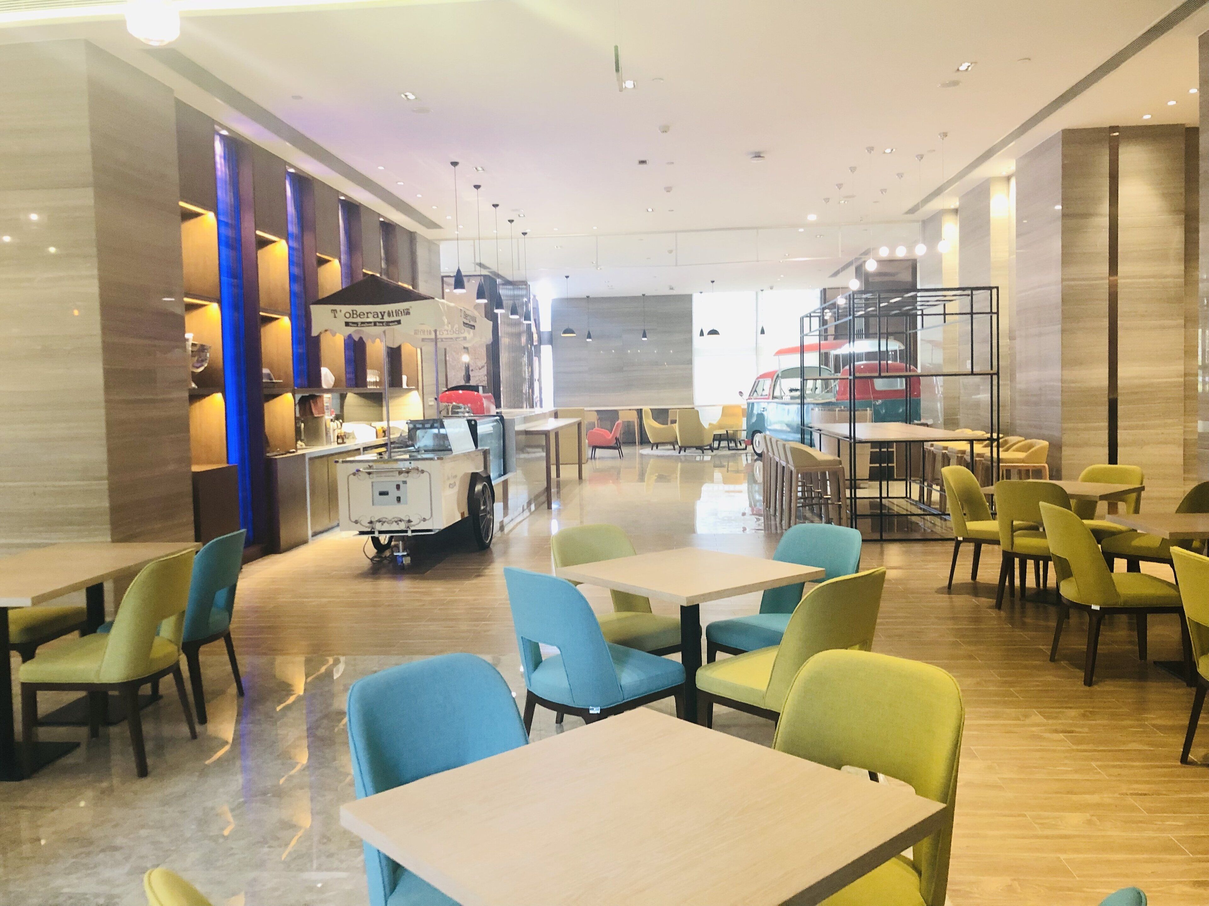 Restaurante moderno com um balcão central de comida, diversas áreas de estar com cadeiras coloridas e um interior limpo e bem iluminado.