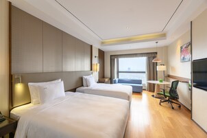 A hotel room with two beds, a desk with a lamp, a chair, a TV, and a window with a view.