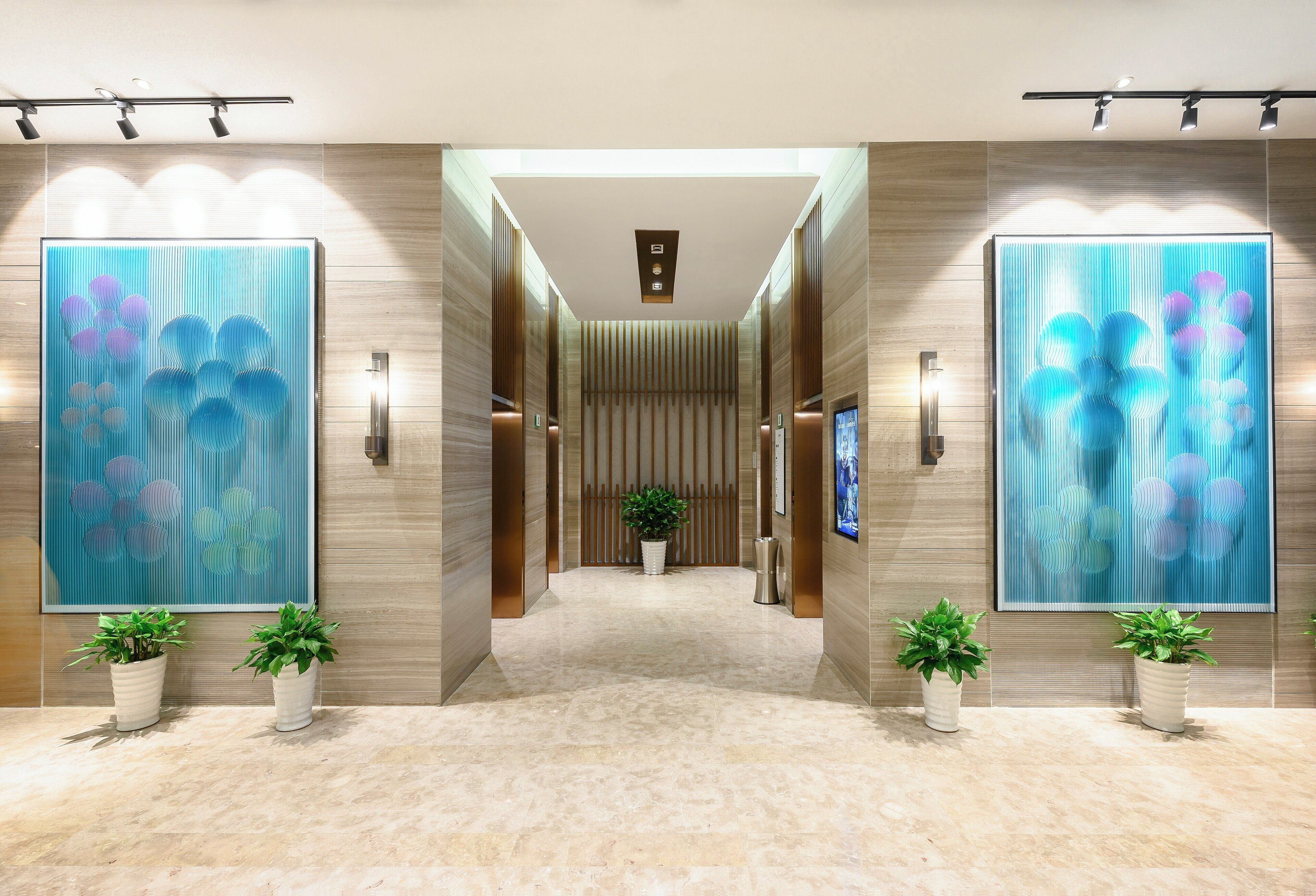 A modern hotel lobby with a central corridor, flanked by large abstract paintings, potted plants, and a row of elevators.