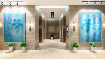 A modern hotel lobby with a central corridor, flanked by large abstract paintings, potted plants, and a row of elevators.