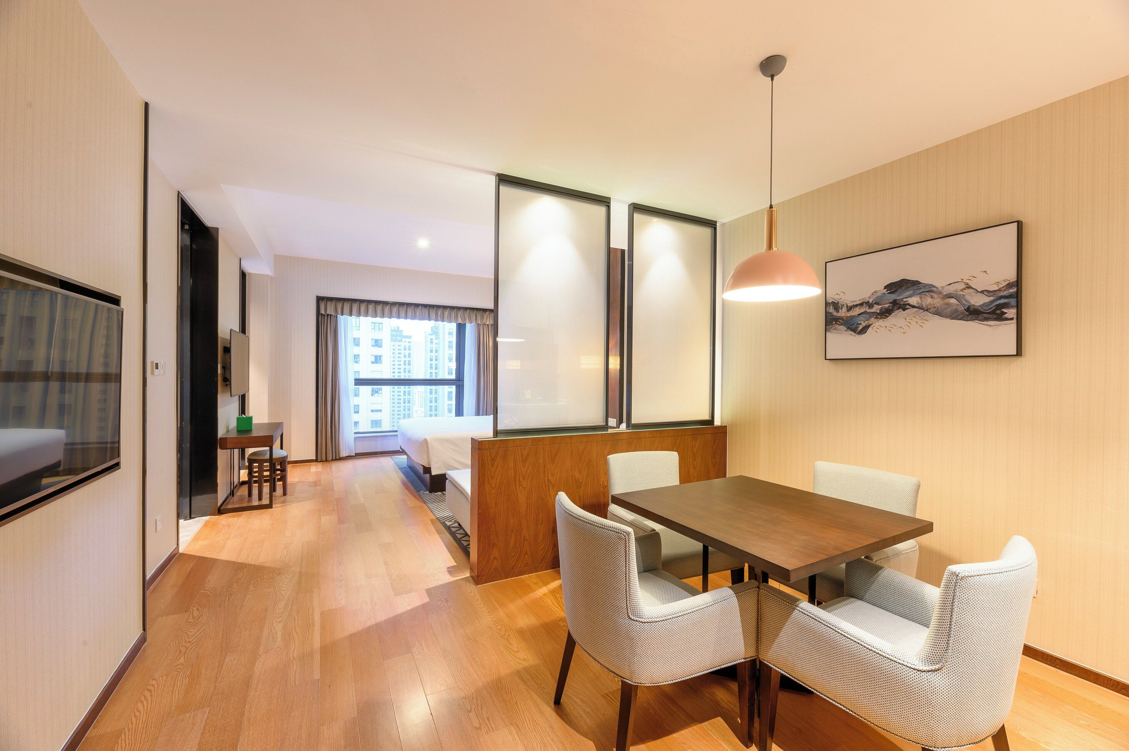 A hotel room with a wooden floor, a dining table with chairs, a bed, a TV, and a large window with curtains.
