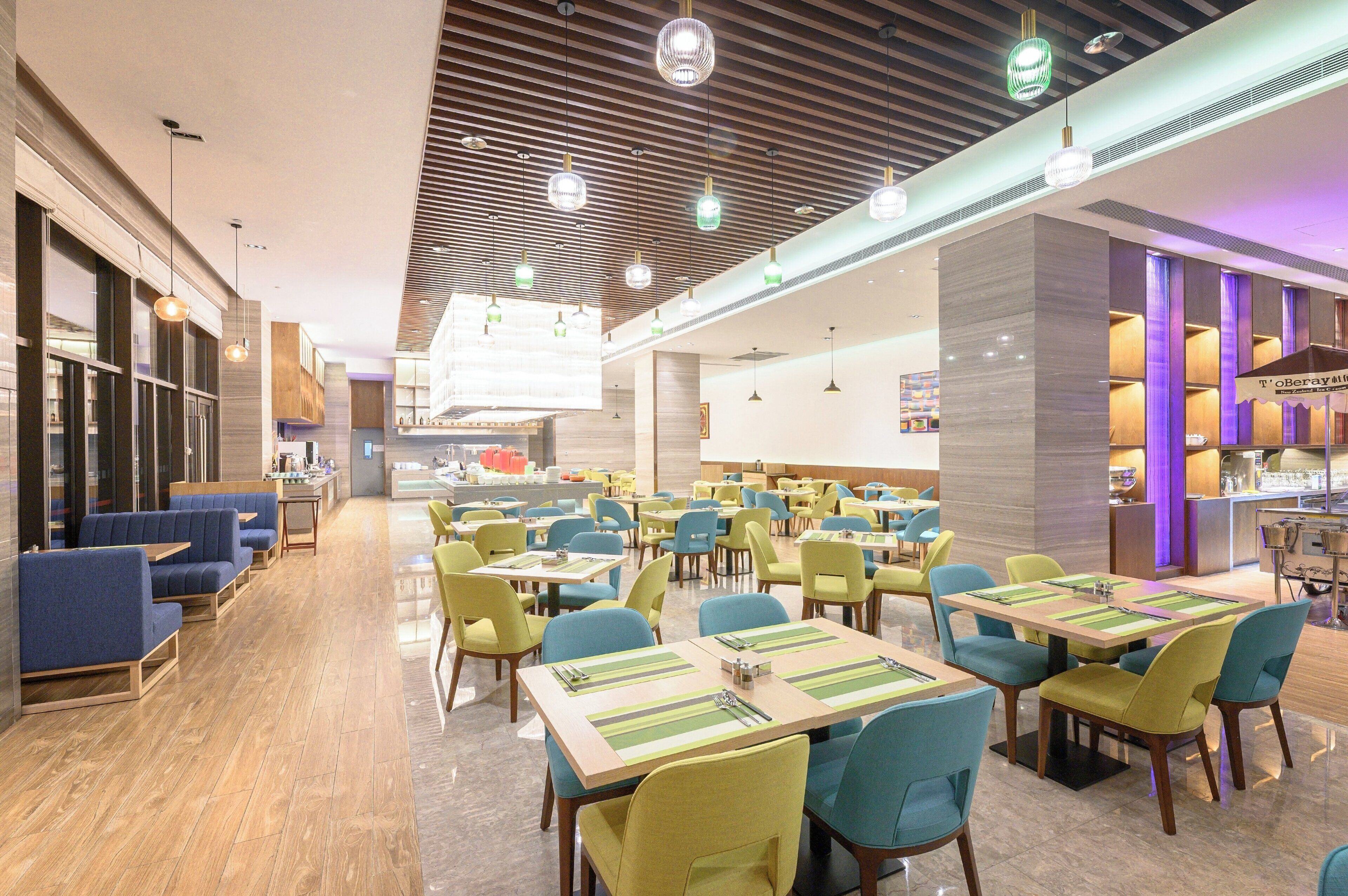 A modern dining area with multiple tables and chairs, a kitchen in the background, and a ceiling with hanging lights.