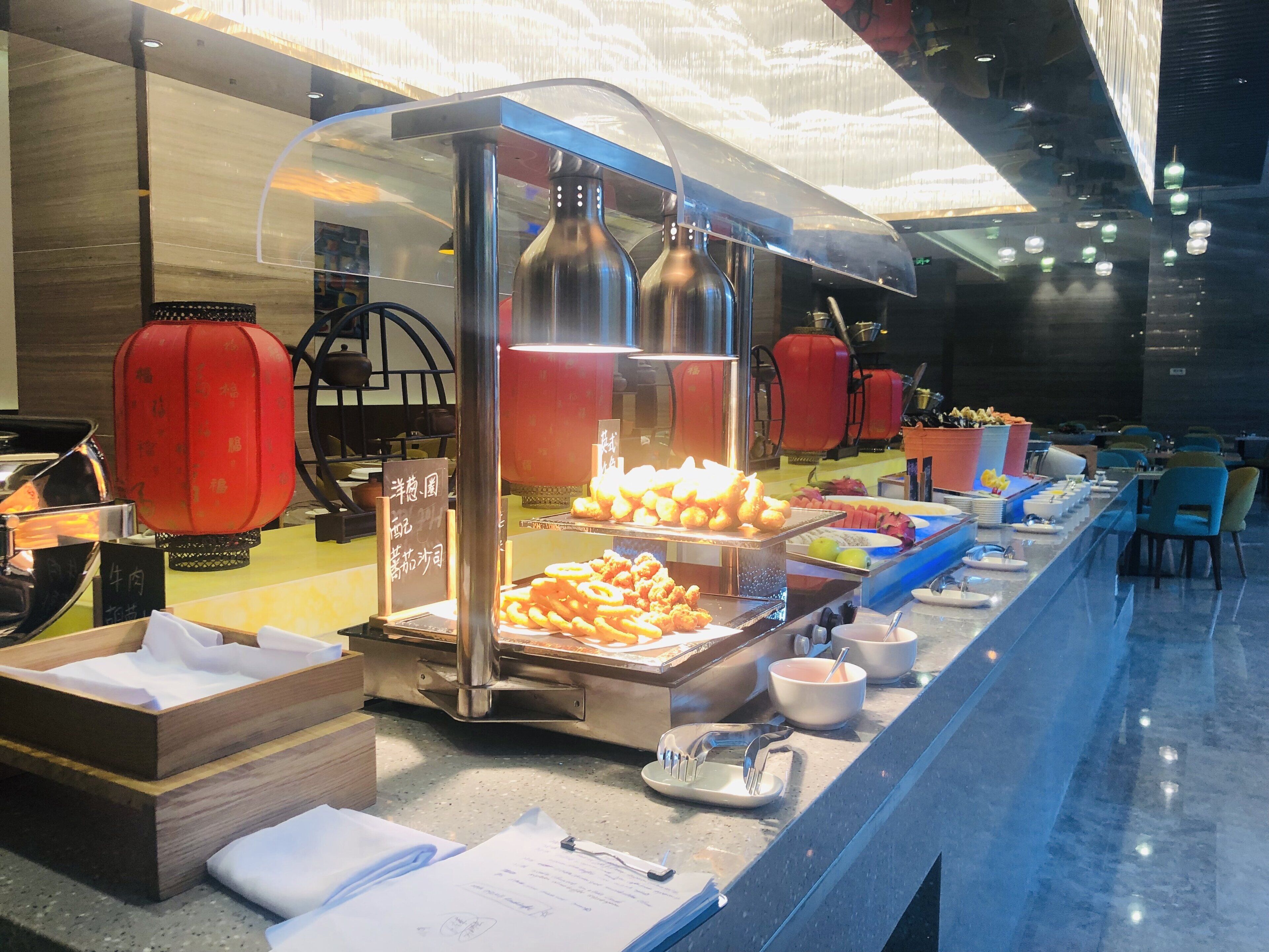 A buffet setup with various food items, including fried snacks and fruits, in a modern dining area with a glass partition and metal serving equipment.