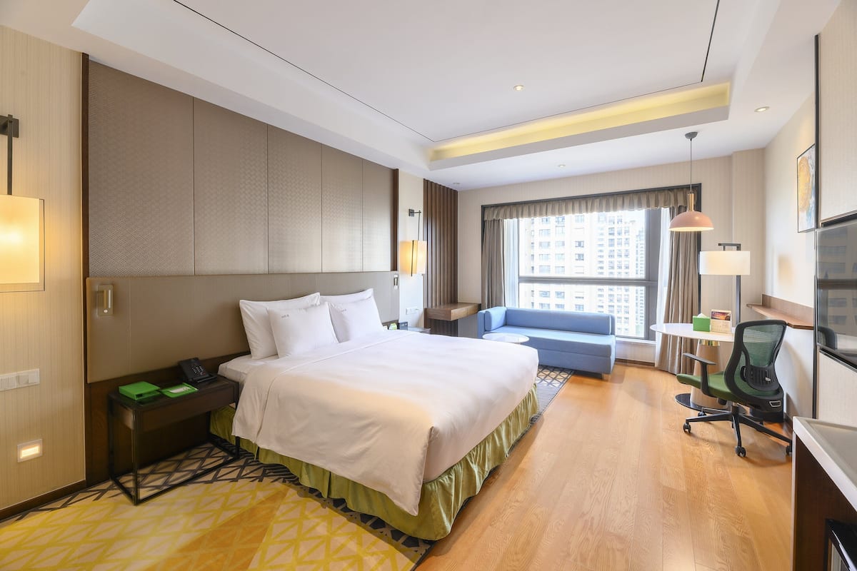 A hotel room with a large bed, a desk with a chair, a sofa, and a window with a view of buildings.