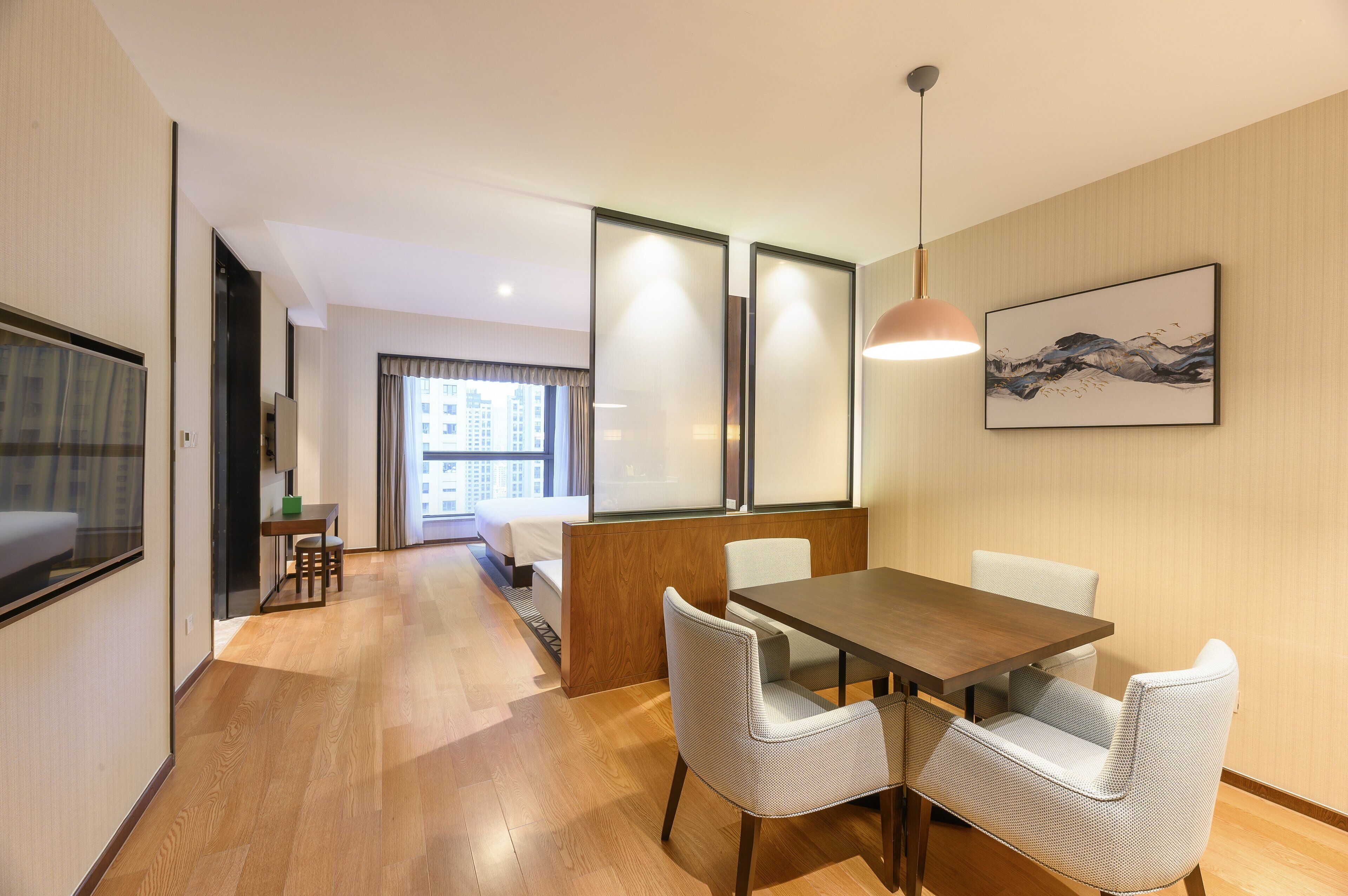 A modern hotel room with a wooden floor, a dining area with a table and chairs, a bed, a window with curtains, and a painting on the wall.