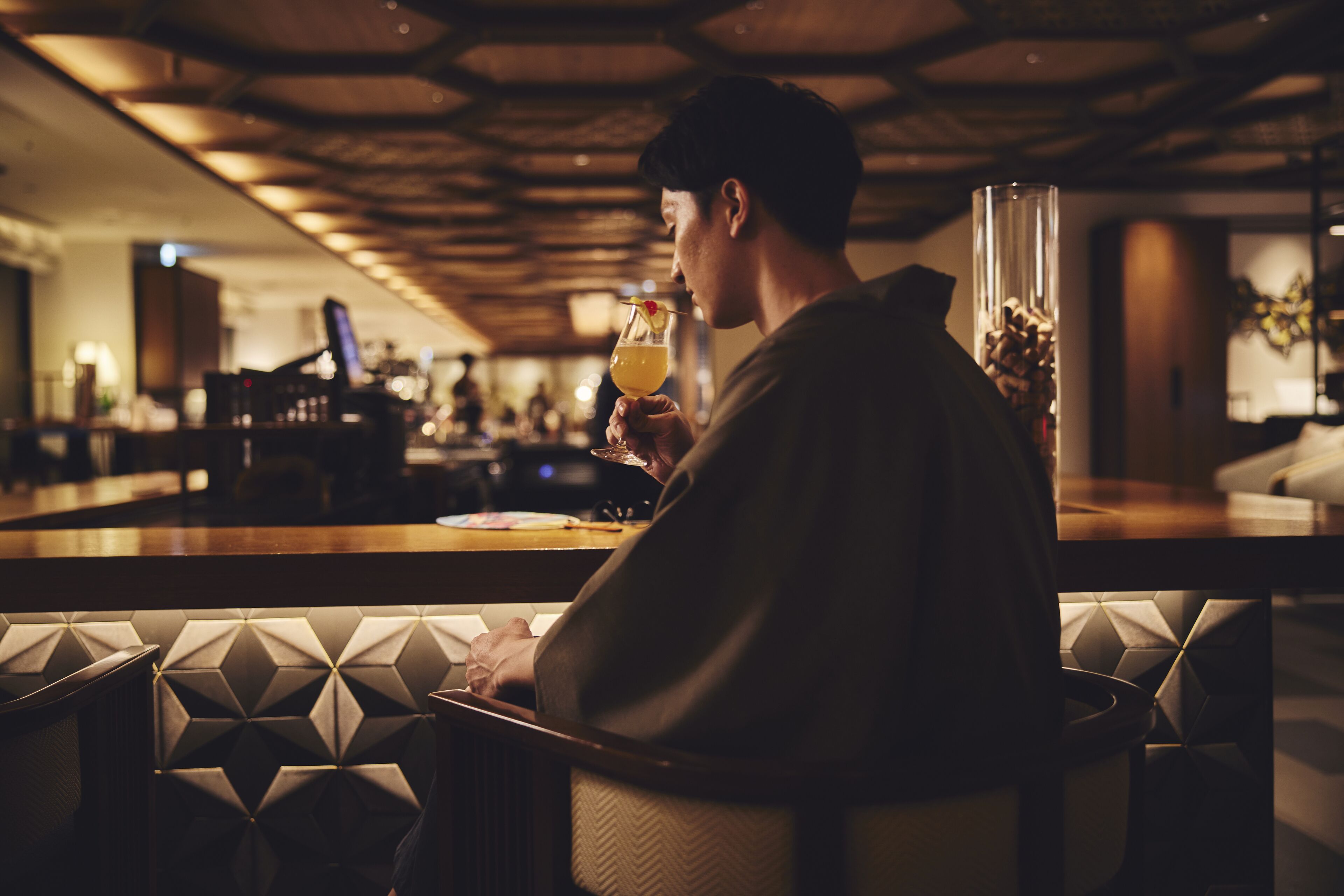 A person seated at a bar with a drink in hand, a bar counter, and a patterned wall in the background.