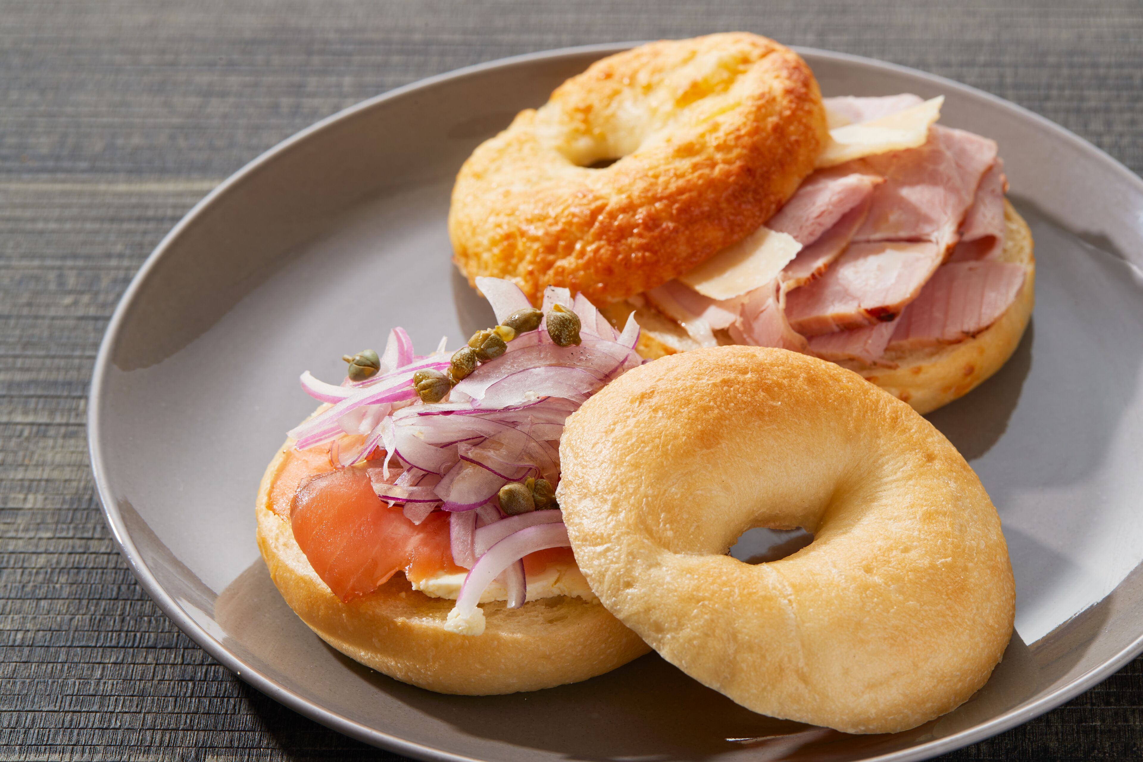 A plate with two bagels, one with smoked salmon and the other with ham, accompanied by red onion and capers.