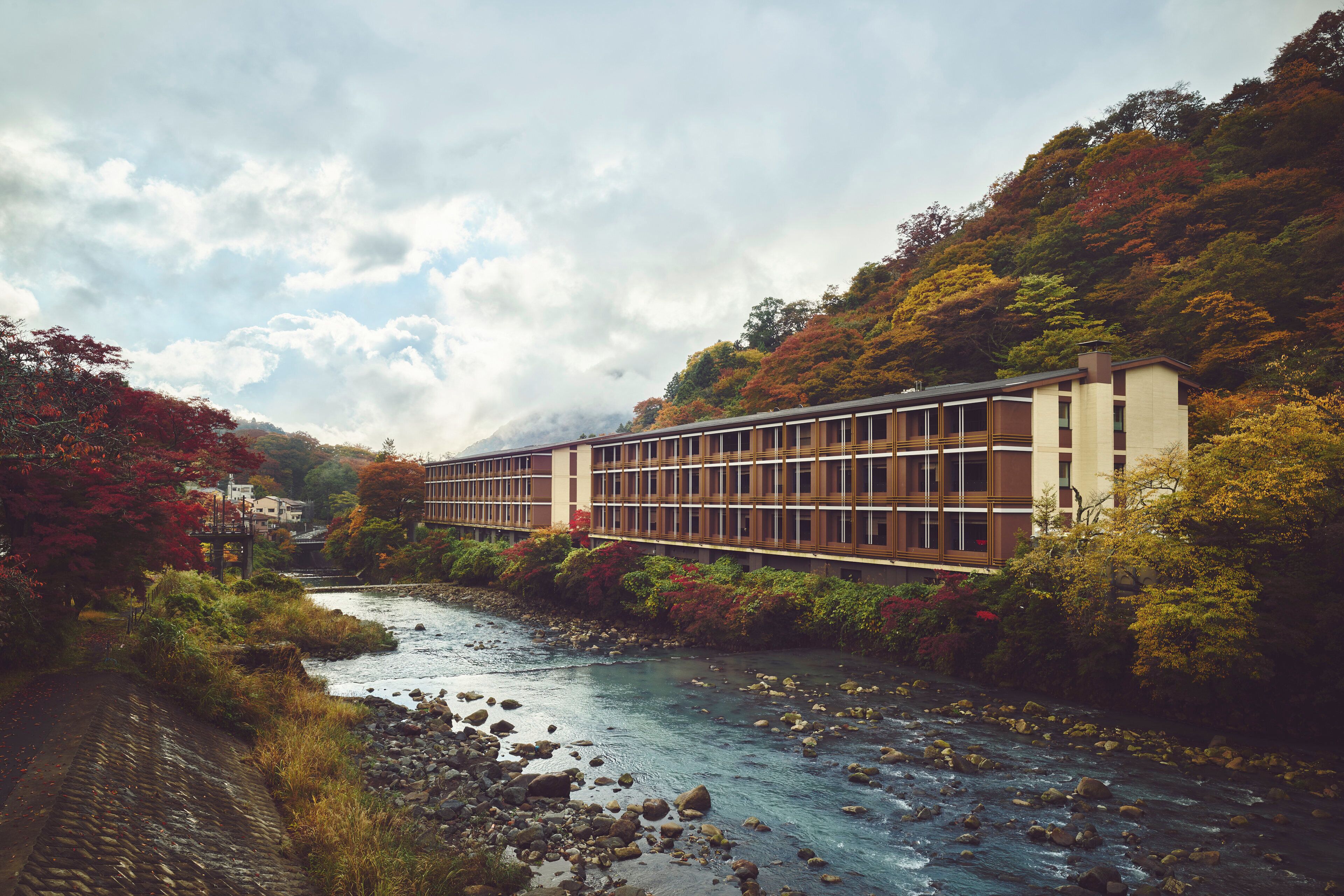 A building with a modern architectural style, a river in the foreground, and a hilly landscape with trees.
