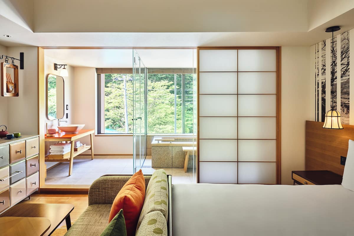 A modern hotel room with a large bed, a sitting area with a sofa and a table, a bathroom with a sink and mirror, and a sliding door leading to a balcony with a view of greenery.