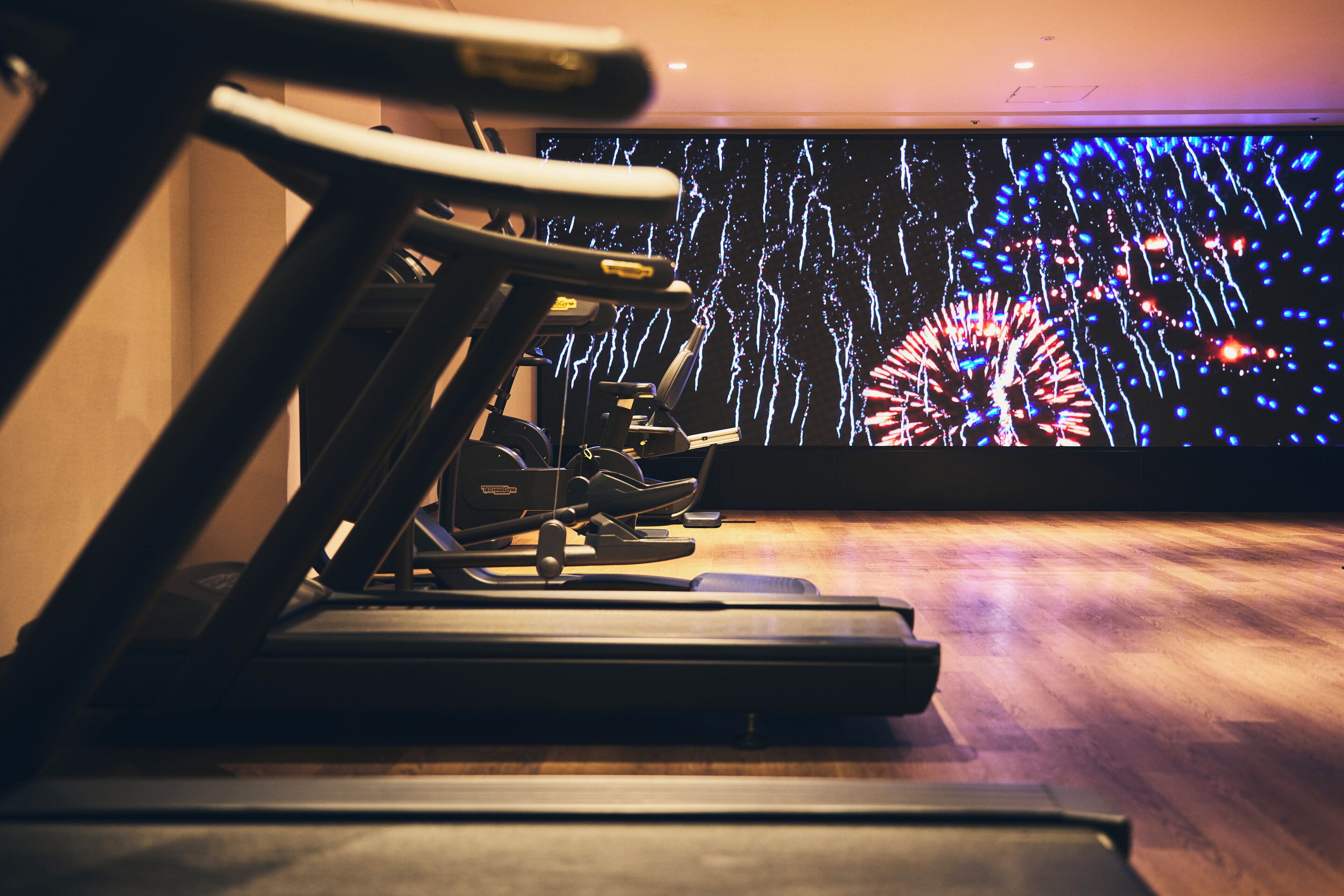 Un tapis de course dans une salle de sport, avec un grand écran affichant des feux d’artifice en arrière-plan.