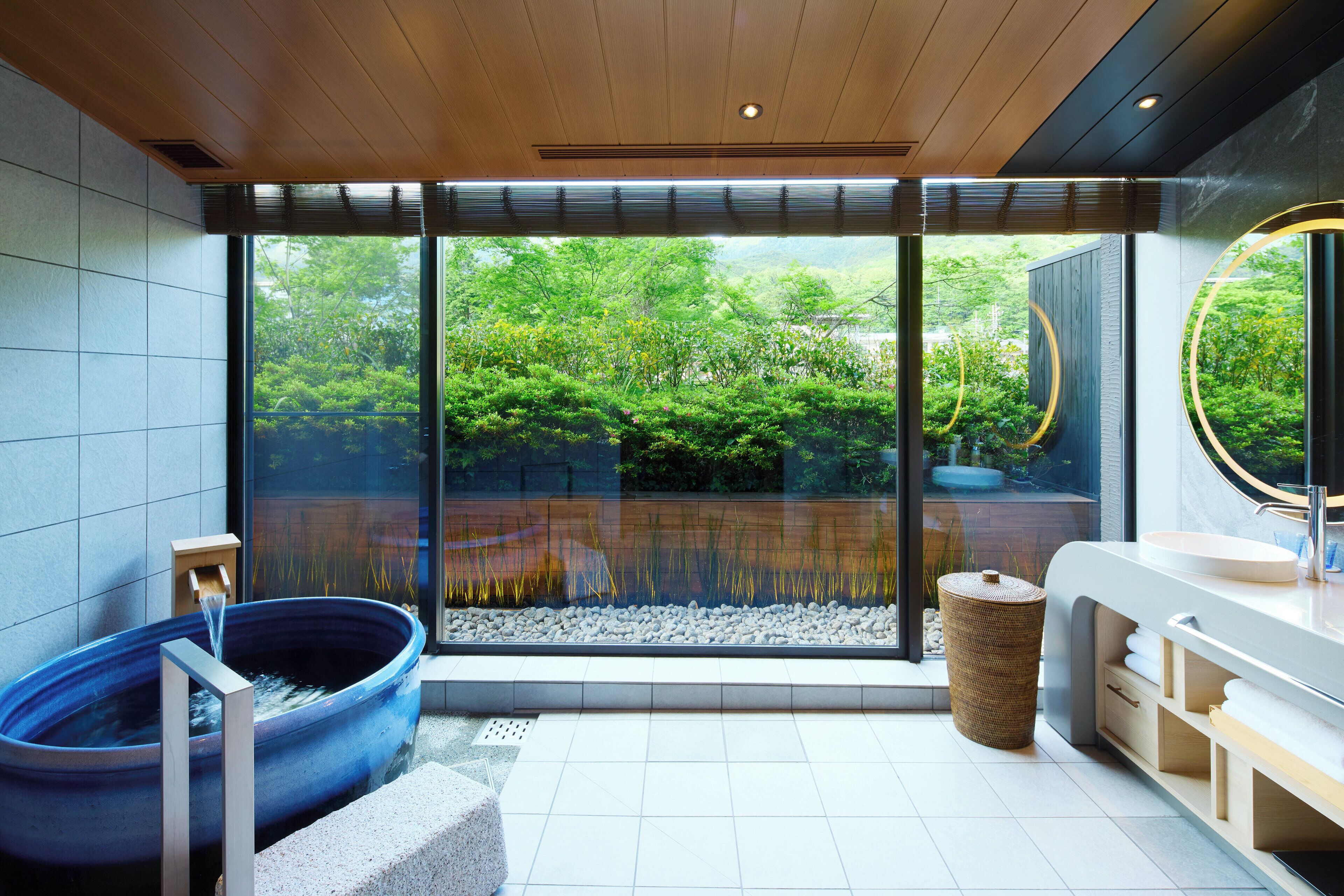A modern bathroom with a large window, a blue bathtub, a sink, and a mirror.