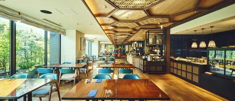 Un restaurante moderno con mesas de madera y sillas azules, una zona de cocina y amplios ventanales con vistas a zonas verdes.