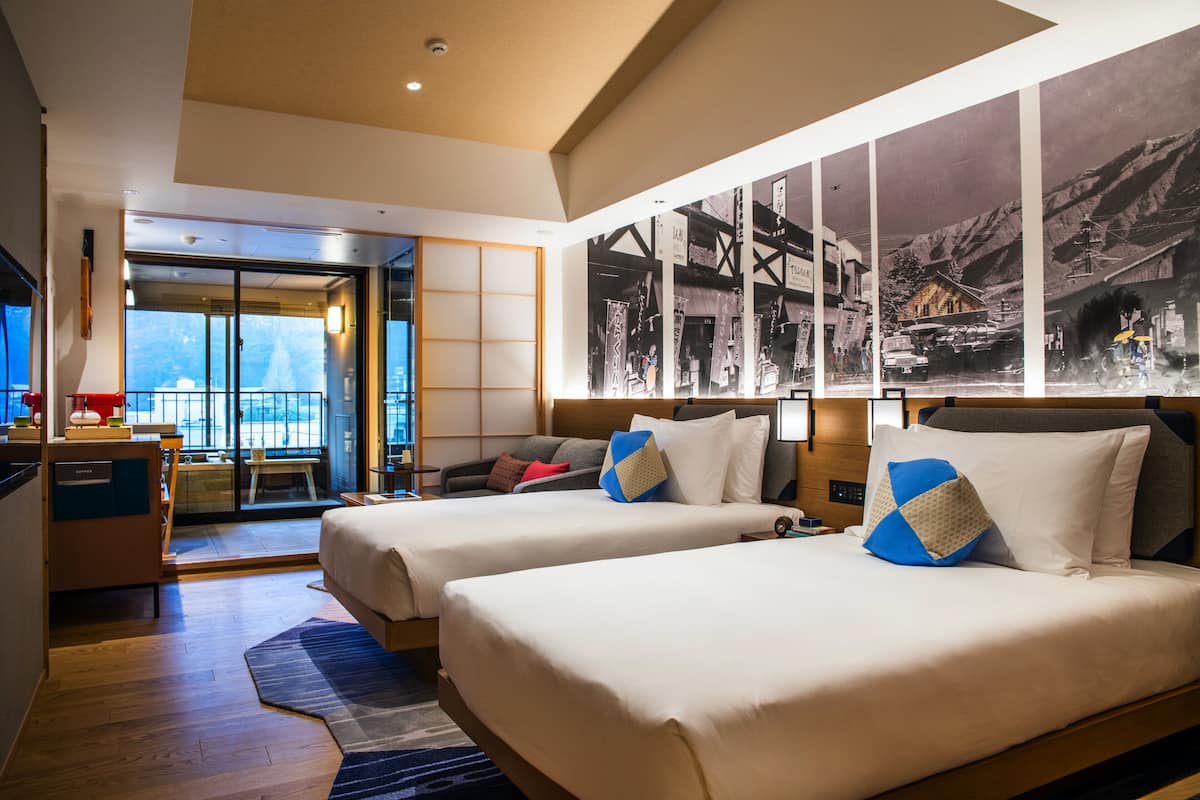 A hotel room with two beds, a desk, a chair, a TV, and a large window with a view.