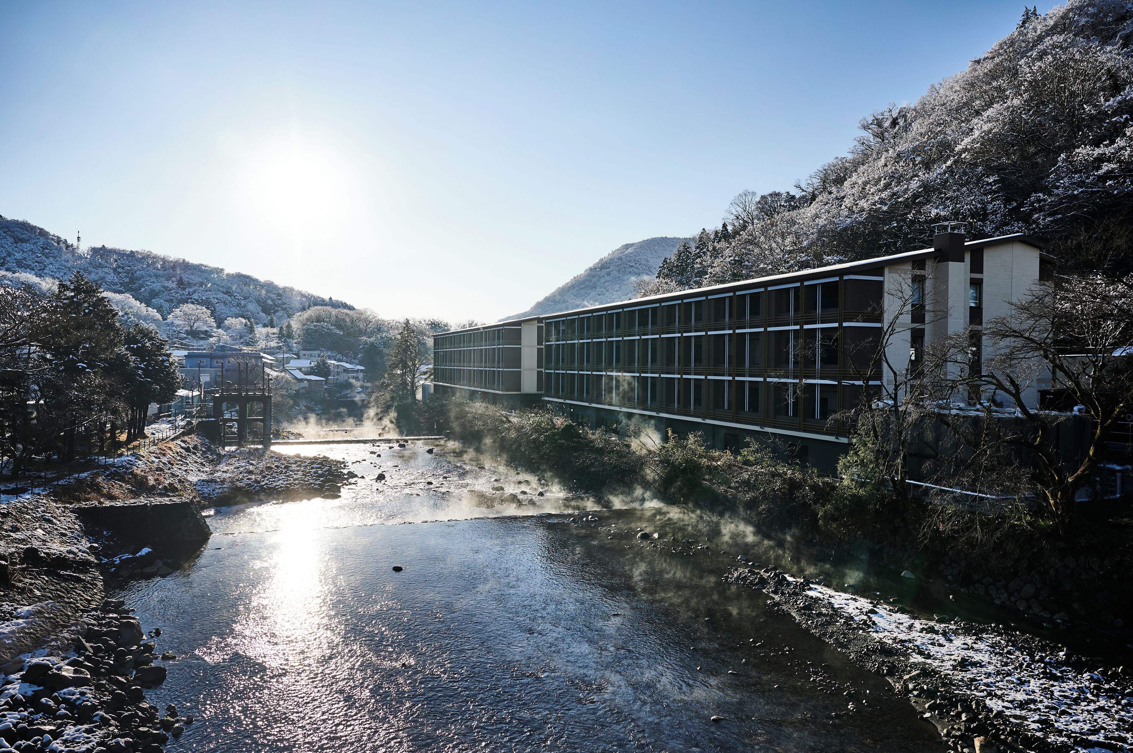 Un edificio moderno con fachada de vidrio que da a un río, rodeado de árboles y colinas cubiertas de nieve.