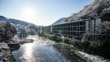 Un edificio moderno con fachada de vidrio que da a un río, rodeado de árboles y colinas cubiertas de nieve.