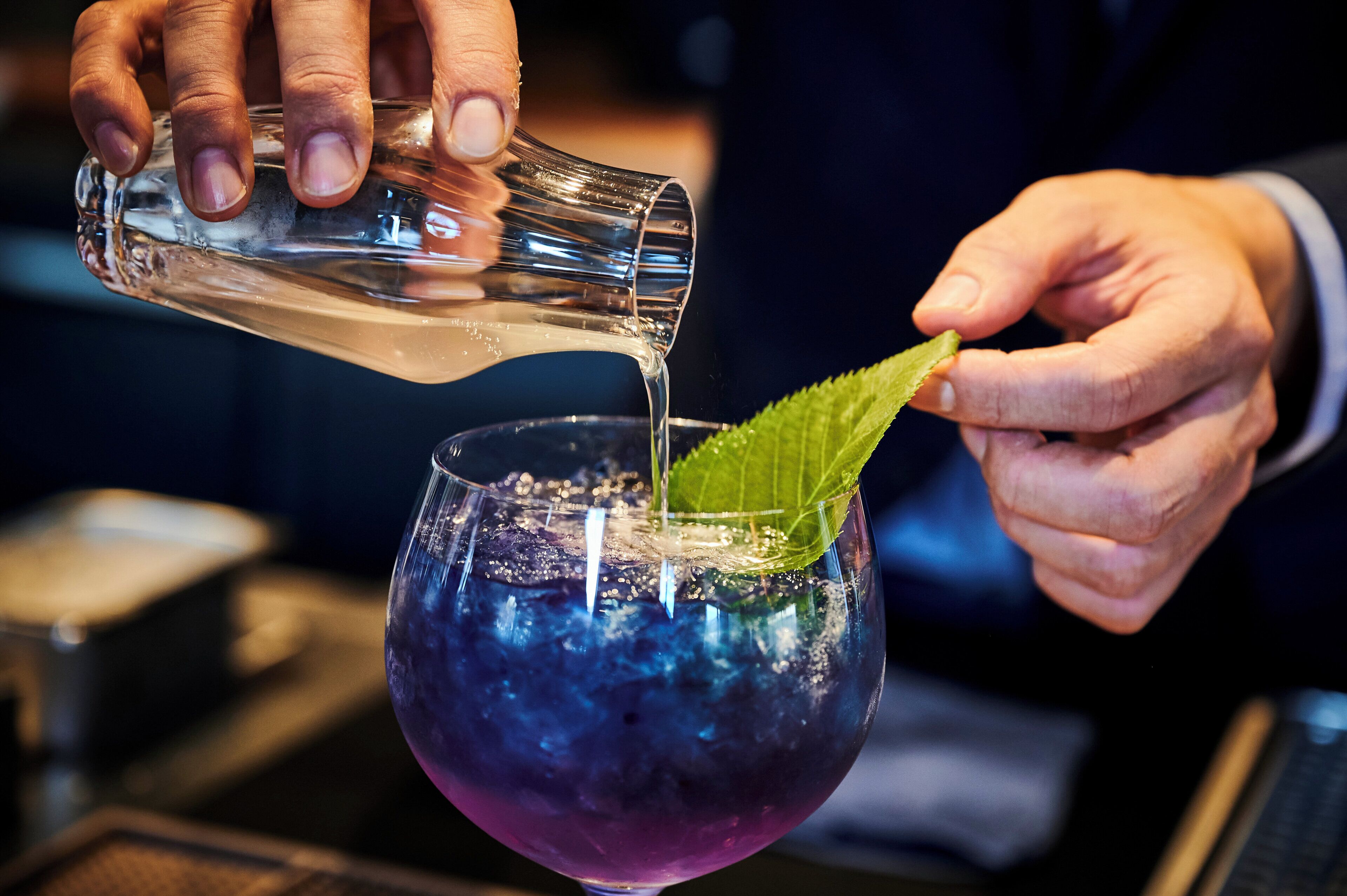 A person pouring a clear liquid into a glass with a purple hue, garnished with a green leaf.