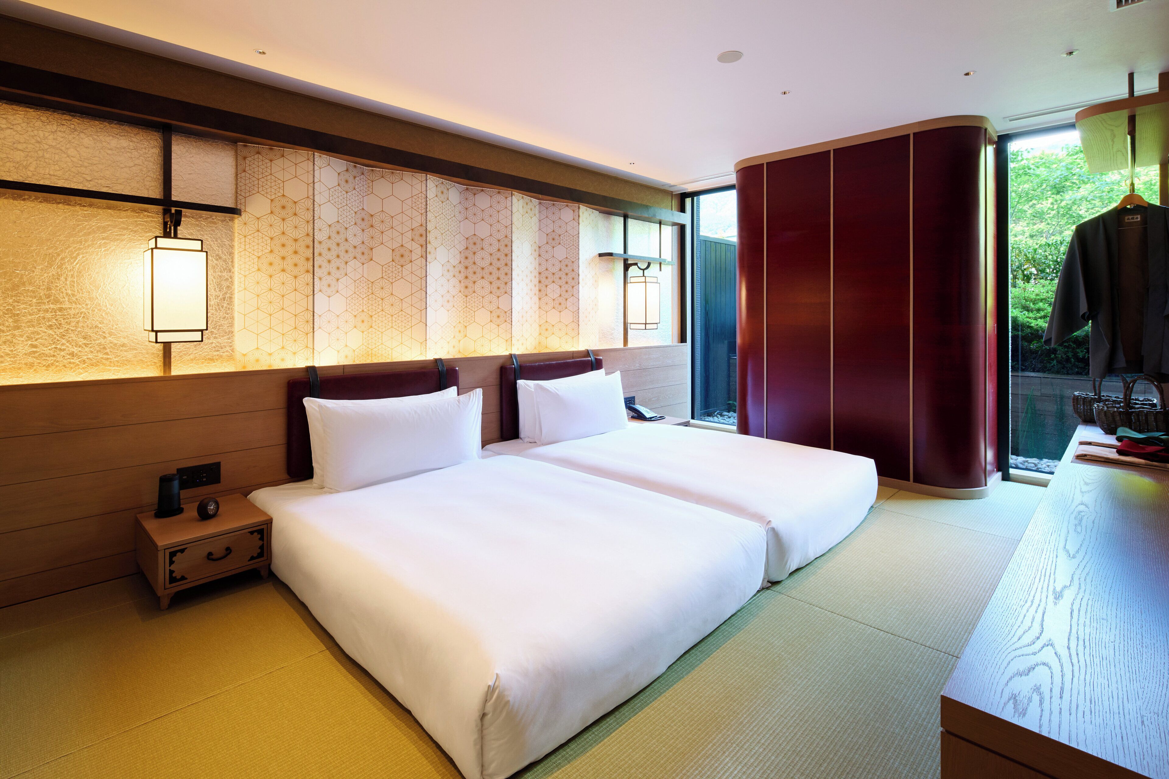 A hotel room with two beds, a wooden nightstand, a traditional Japanese-style wall, and a view of greenery through a sliding door.
