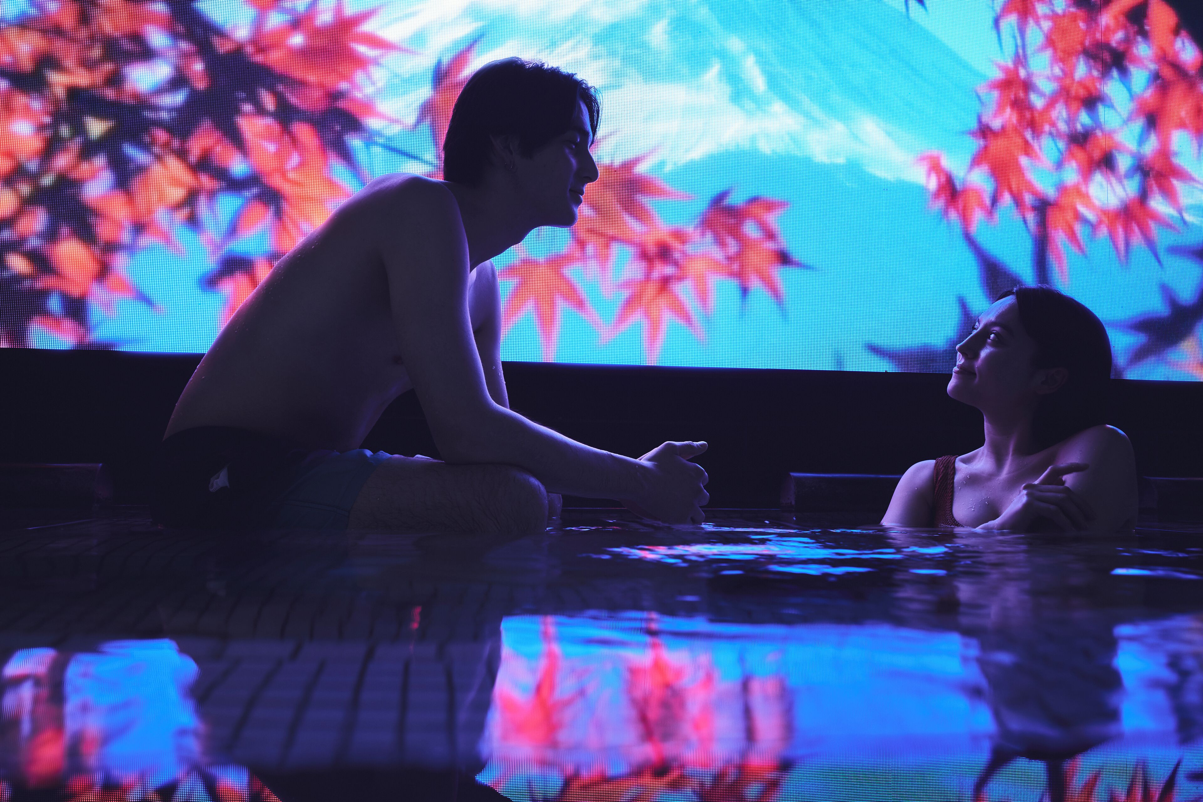 Una persona sentada junto a una piscina, con una gran pantalla digital que muestra hojas de colores en el fondo.