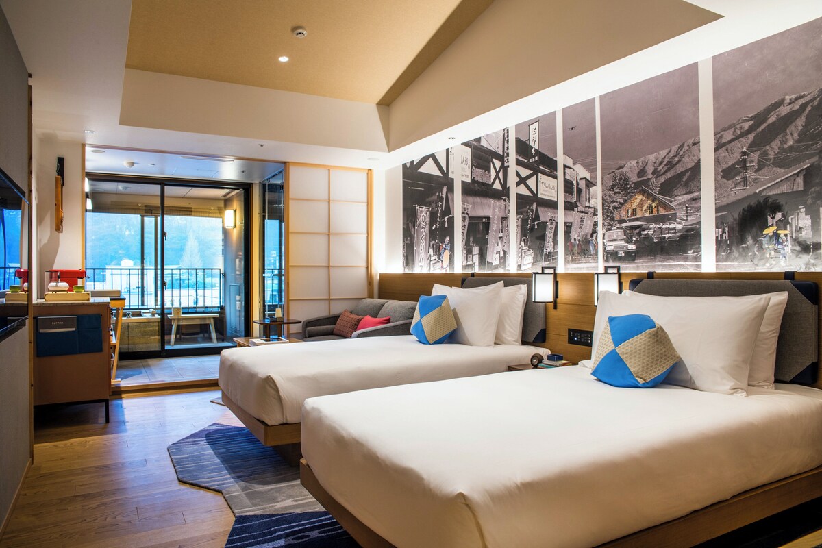 Habitación de hotel con dos camas, un escritorio, una silla, un televisor y un ventanal con vistas.
