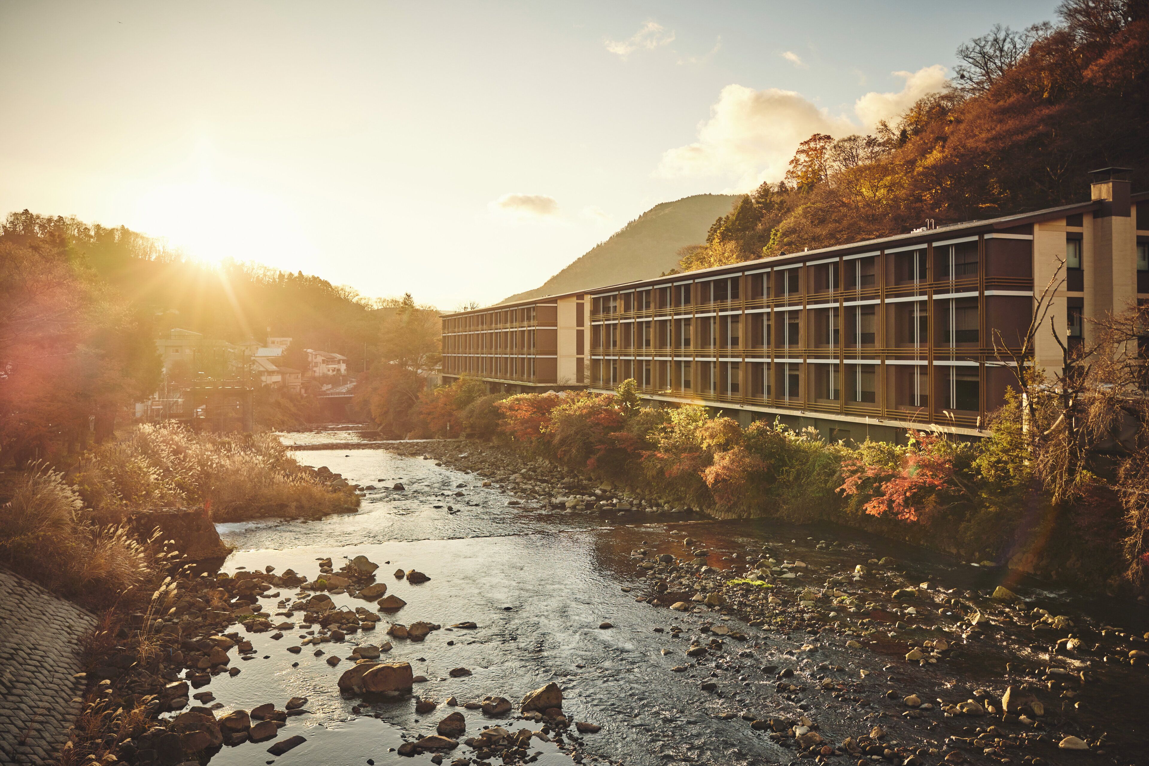A modern building with a long facade, a river in the foreground, and a hilly landscape in the background.
