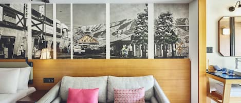 Habitación de hotel moderna con un sofá gris, dos almohadas, una mesita de madera y un mural grande con un paisaje montañoso.