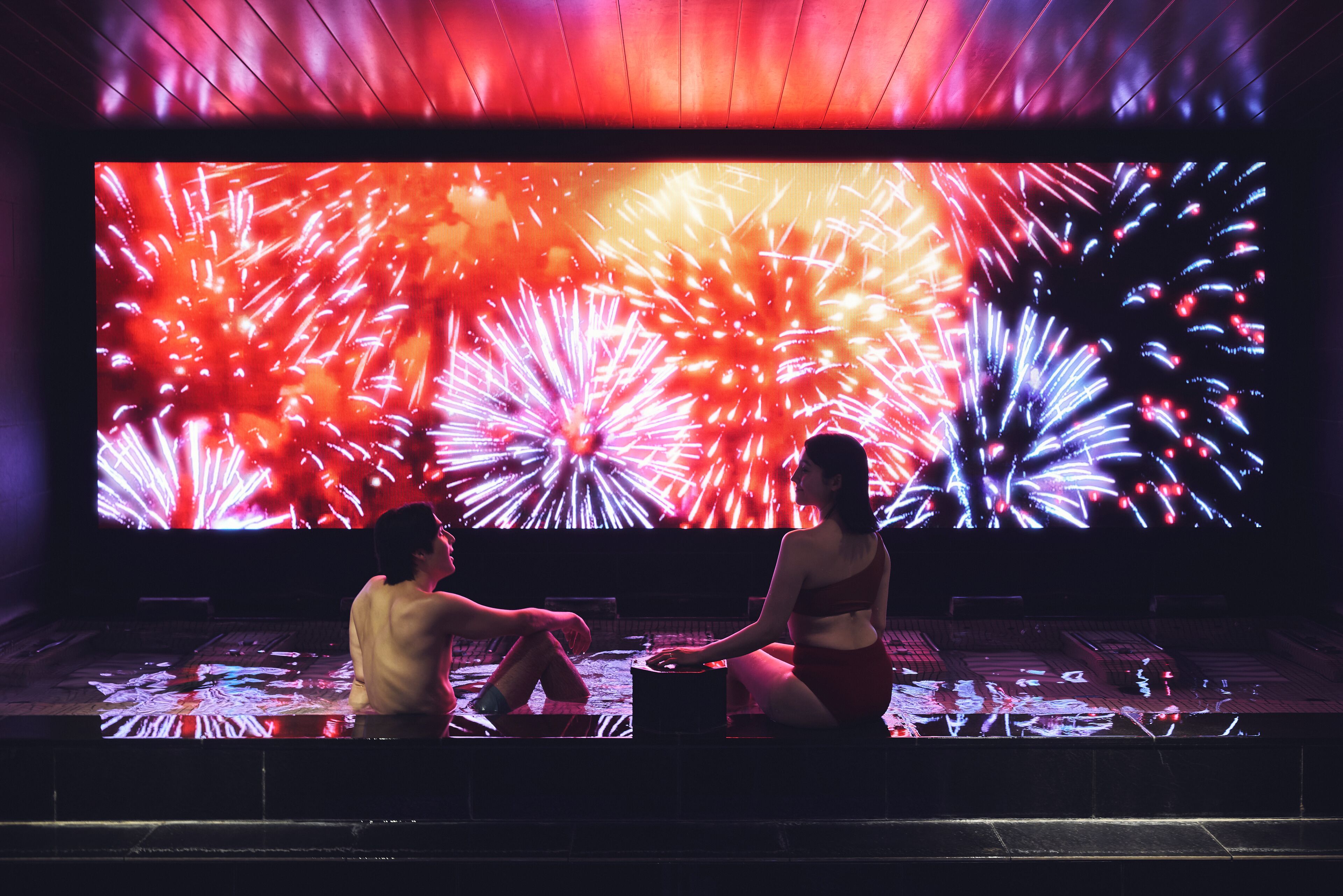 Una pareja sentada en un banco mirando una pantalla grande que muestra fuegos artificiales.