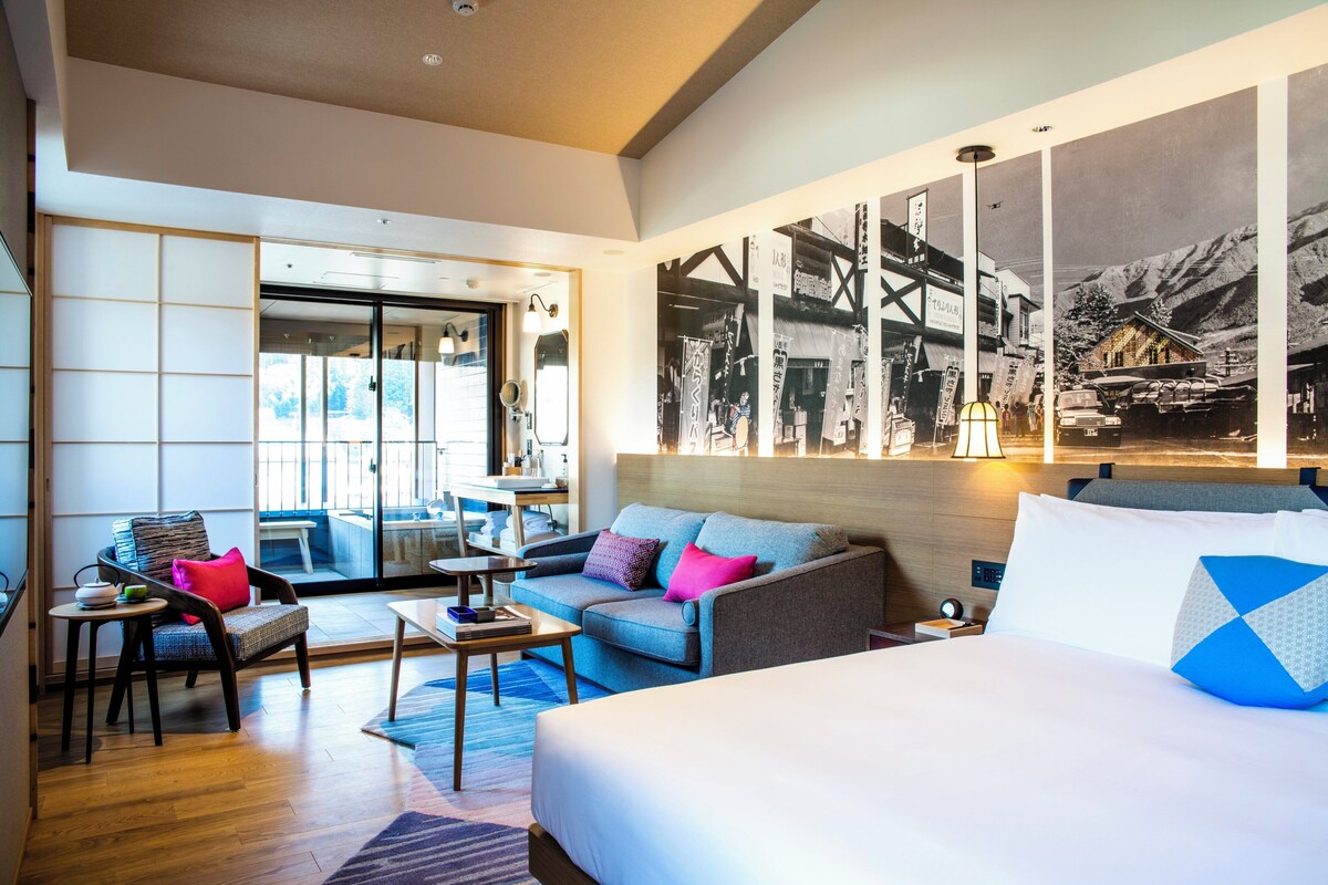 Habitación de hotel con cama, sofá, sillón, mesita y vistas al exterior.