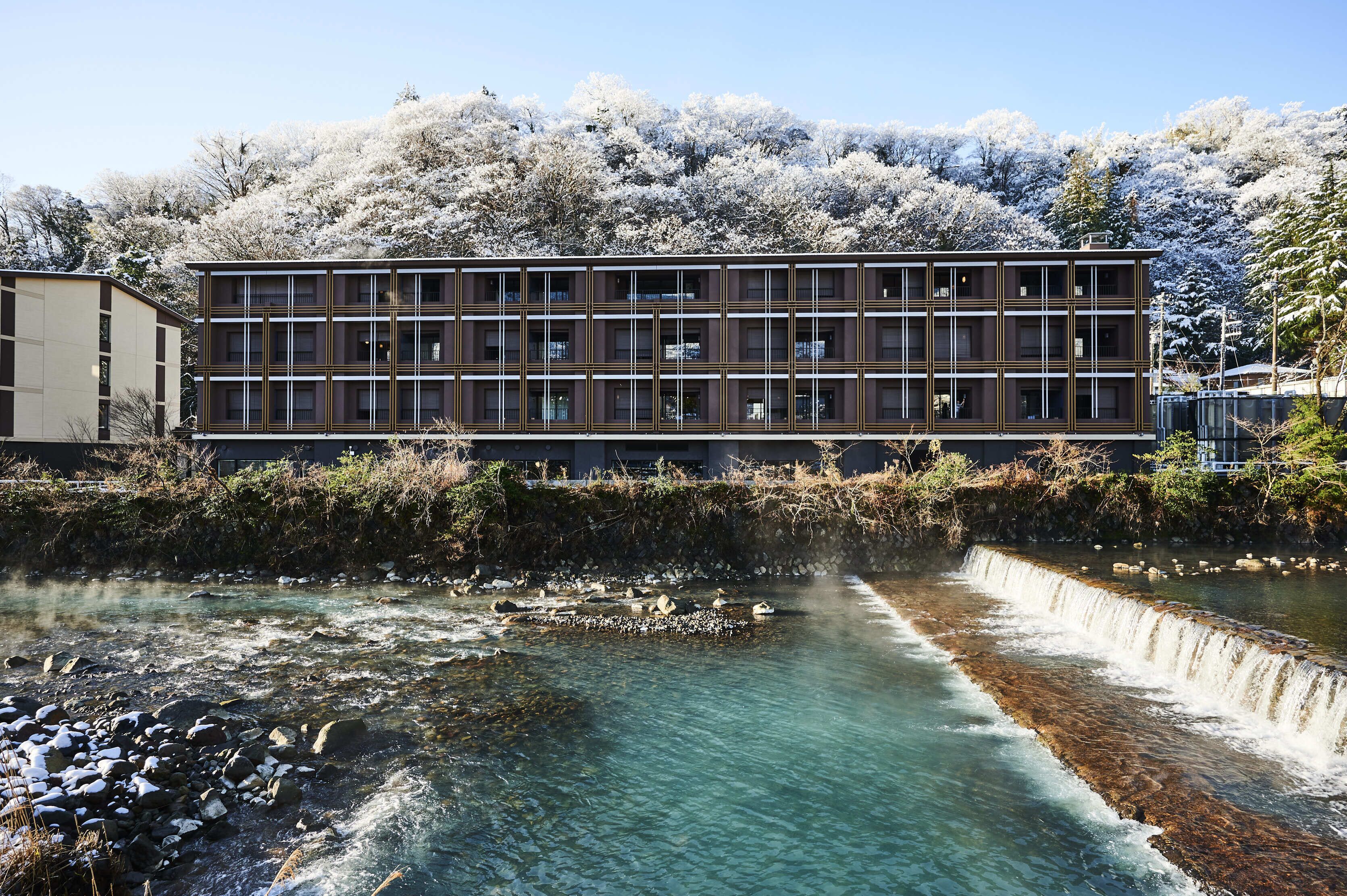 A modern building with a multi-story structure, a river in front, and a snowy hillside in the background.