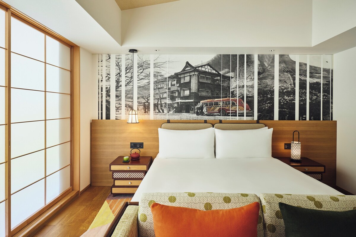 Habitación de hotel con una cama grande, cabecera de madera, mesita de noche con lámpara, televisor montado en la pared y un panel corredizo shoji.