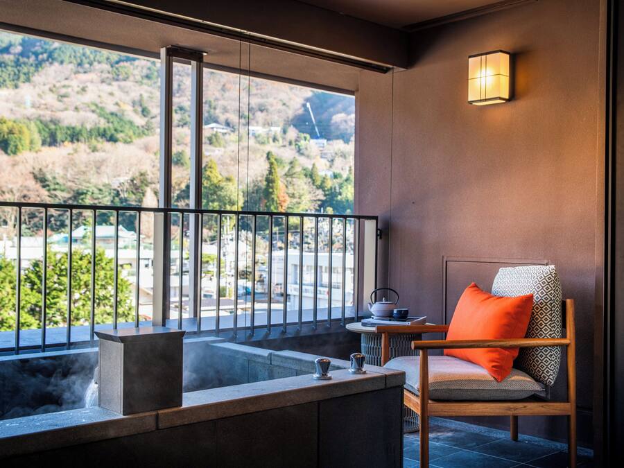 A balcony with a view of a mountainous landscape, a seating area with a chair and a table, and a wall-mounted light fixture.