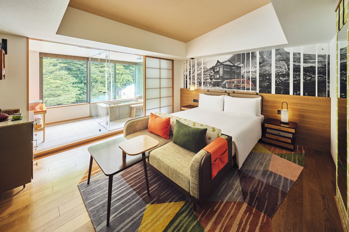 A modern hotel room with a large bed, a sofa, a small table, and a view of greenery through the window.