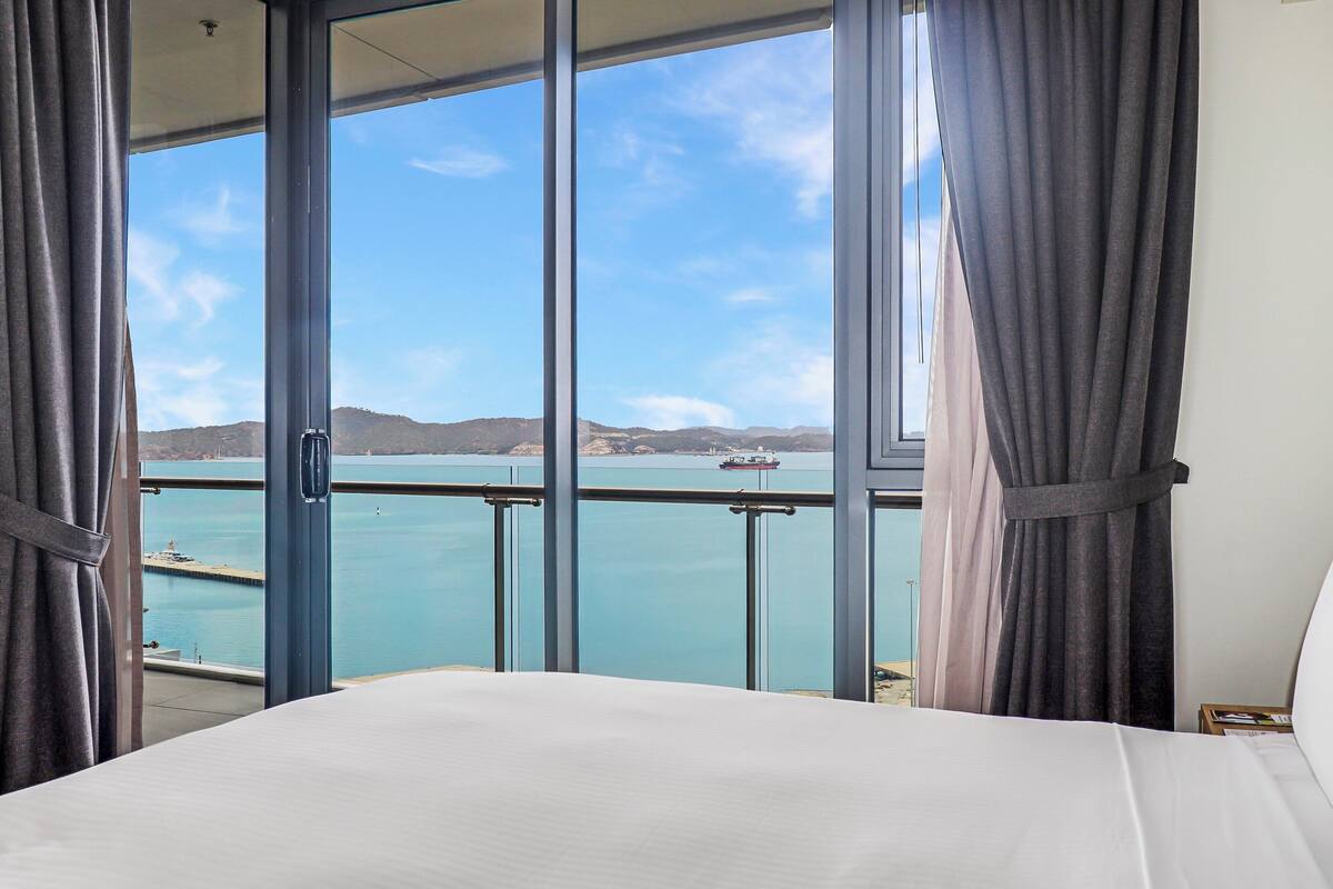 A hotel room with a large window offering a view of a calm sea and distant hills.