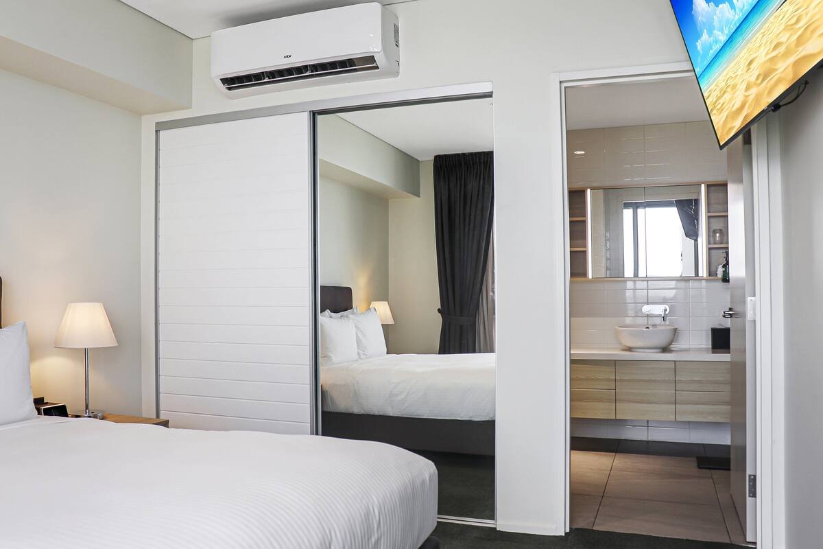A hotel room with a bed, bedside table, lamp, air conditioning unit, and a TV screen displaying a beach scene.