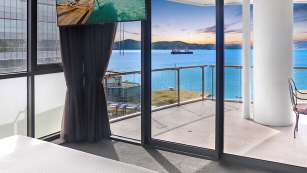 Una habitación con un amplio ventanal que da a una extensión de agua, un balcón y vistas a un atardecer.