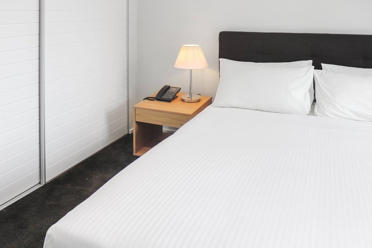 A neatly made bed with white linens, a bedside lamp, and a telephone on a wooden nightstand.