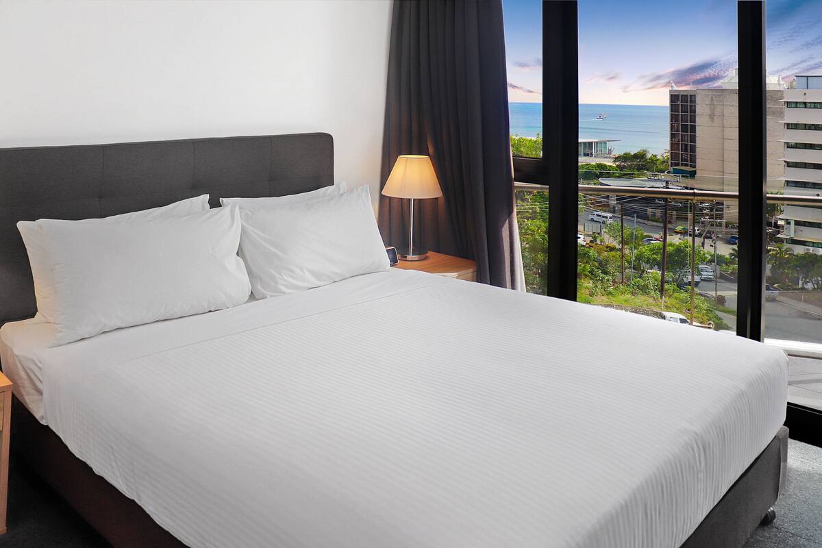 A hotel room with a large bed, a nightstand with a lamp, and a view of the ocean and cityscape through large windows.