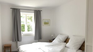 Une chambre à coucher avec un lit, une table de chevet, une fenêtre avec des rideaux, un radiateur et un petit tabouret.